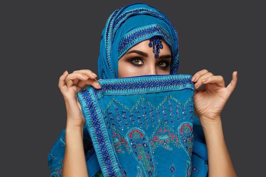 Close-up portrait of a white-skinned girl with beautiful smoky eyes wearing an elegant blue hijab decorated with sequins and jewelry. She is covering her face with a shawl and looking at the camera on a dark background. Human emotions, facial expression concept. Trendy colors. Arabic style.