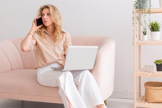 A blonde business woman, in light clothes, sits on a light pink sofa, with a gray open laptop on her knees, calls on a pink smartphone