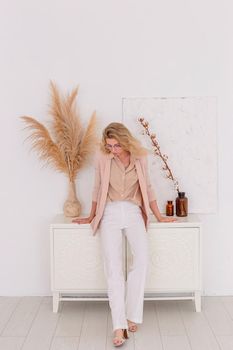 a beautiful slender woman of 30-40 years old with blond hair is standing in a light pink jacket and white trousers in the interior of a white room, near a chest of drawers. Copy space. Vertical