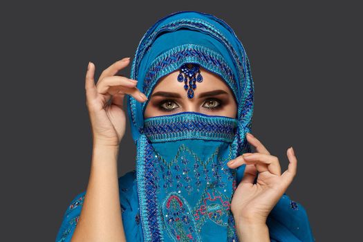 Close-up portrait of a lovely young woman with beautiful smoky eyes wearing a blue hijab decorated with sequins and jewelry. She is gesticulating and looking at the camera on a dark background. Human emotions, facial expression concept. Trendy colors. Arabic style.