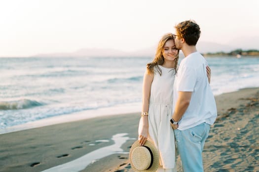 Man hugs and kisses woman on the forehead while standing on the beach by the sea. High quality photo