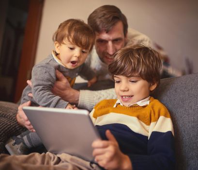 Lets see what your brothers doing. an adorable little boy using a tablet on the sofa at home while his father and younger brother look on