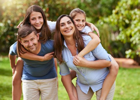 Alright where are we going. Portrait of a cheerful young mother and father giving their daughters a piggyback ride outside in a park during the day