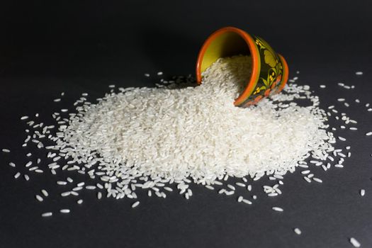 Scattered rice uncooked grains with painted wooden cup on a black background.
