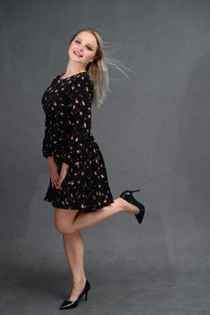 Full-length portrait of a pretty young smiling woman on a gray background in a black dress with long straight hair. Standing straight at full height, showing emotions, talking in different poses.