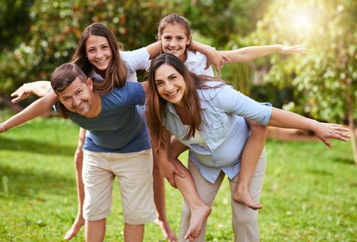 Come fly with us. Portrait of a cheerful young mother and father giving their daughters a piggyback ride outside in a park during the day