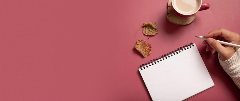 Table desk with notepad and female hand wit pencil and fall leaves on burgundy colored background. Autumn flat lay composition with copy space. Banner.
