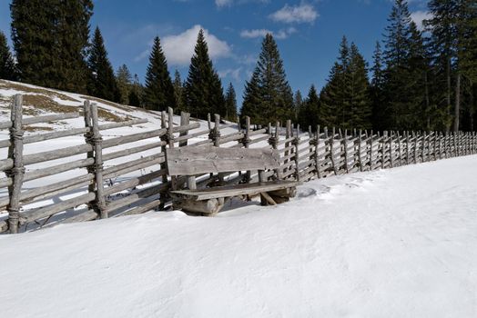 In front of an old wooden fence stands a massive heavy wooden bench on a snowy meadow. The warming sun's rays and the blue sky invite you to rest. Some large fir trees can be seen in the background.