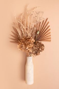 Bouquet of beige dried flowers, grass and leaves in a glass vase on beige background top view.
