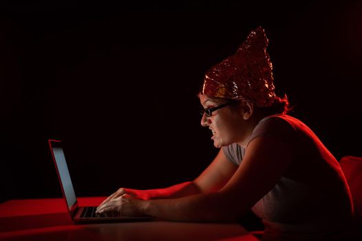 Paranoid caucasian woman typing on a computer wearing a tinfoil cap. Conspiracy theory