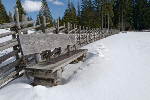 In front of an old wooden fence stands a massive heavy wooden bench on a snowy meadow. The warming sun's rays and the blue sky invite you to rest. Some large fir trees can be seen in the background.