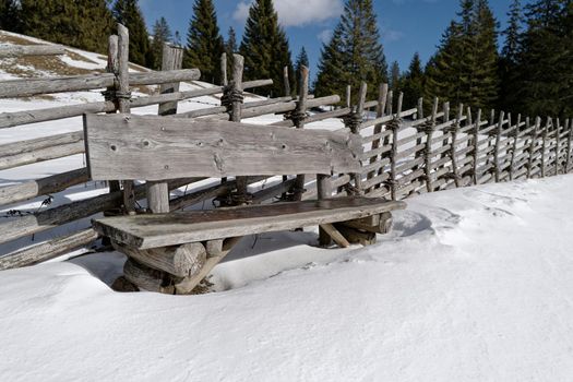 In front of an old wooden fence stands a massive heavy wooden bench on a snowy meadow. The warming sun's rays and the blue sky invite you to rest. Some large fir trees can be seen in the background.