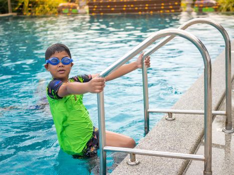 The boy wearing swimming goggles on blue pool ladder green swimsuit