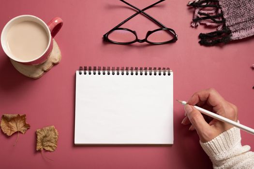 Table desk with notepad and female hand wit pencil and fall leaves on burgundy colored background. Autumn flat lay composition with copy space.