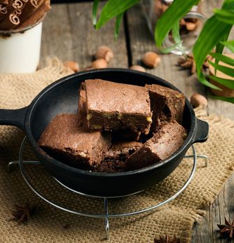 Baked pieces of chocolate brownie cake with walnuts in a black metal frying pan on a wooden table, top view