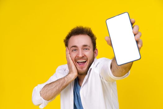 Great offer. Young happy man holding smartphone showing a white empty screen and exciting to win isolated over yellow background, celebrating success. Product placement. Mobile App Advertisement.