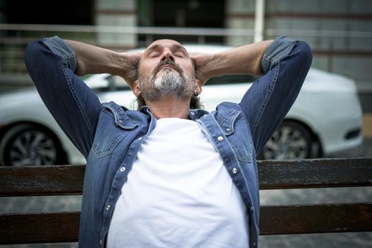 Relaxed, enjoying life handsome middle age man sits leaned back with hands behind his head on the bench outdoors at urban city background wearing denim shirt and white t-shirt. Travel concept.