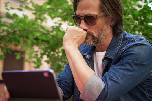 Puzzled up, sad middle aged man working outdoors using digital tablet. Handsome freelancer traveling man not happy checking work or social media sitting at city street cafe.