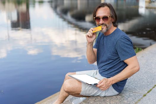 Biting credit, debit card excited freelancer man sitting outdoors next to river working using digital tablet pc, shopping online or paying online banking. E-banking concept. Financial concept.