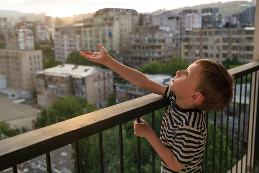 A 6-year-old boy climbs the dangerous high railing of a balcony unsupervised. City in the evening at sunset. Child at home alone. Risky child behavior. Deadly accidents. Altitude, danger