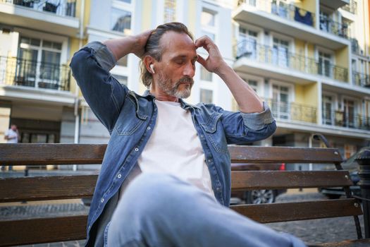 Handsome middle aged man sits on the bench listening music using wireless earphones, with hands lifted up fixing his long hair and eyes shut at old town background enjoying free time.