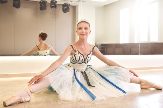 young ballerina practicing ballet poses sitting against the mirror in the dance studio.