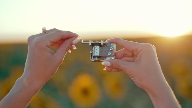 Female hands rotating gears of old music box mechanism. Lady turning the lever of retro small metallic carillon. Woman in sunflowers field listening to music which playing. High quality photo