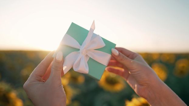 Woman holding blue gift box with bow on sunflowers field background with sun flares. Present, bonus, offer concept. Only hands. High quality photo