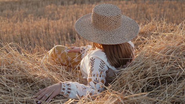 Pretty woman in straw hat and embroidered blouse smiling lying on hay in countryside at sunset. Rural nature, haystack, vacation, relax and harvest concept. High quality photo