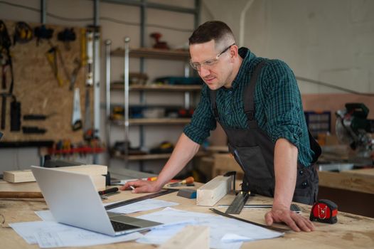 Portrait of a carpenter in protective glasses and work overalls uses a laptop in a workshop