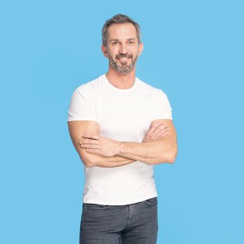 Handsome middle aged grey haired man standing with arms folded happy looking at camera wearing white t-shirt and jeans isolated on blue background. Mature fit man, healthy lifestyle concept.
