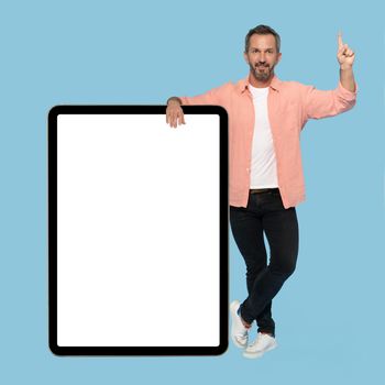 Pointing finger up middle aged grey haired man leaned on big digital tablet with white screen, happy smiling on camera wearing peach shirt isolated on blue background.