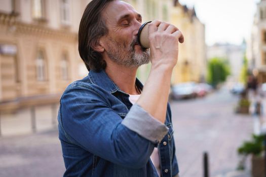 Enjoying his coffee handsome middle aged man with grey beard in denim standing in the city drink take away coffee from paper cup. Handsome man traveling enjoying free time. Business travel concept.