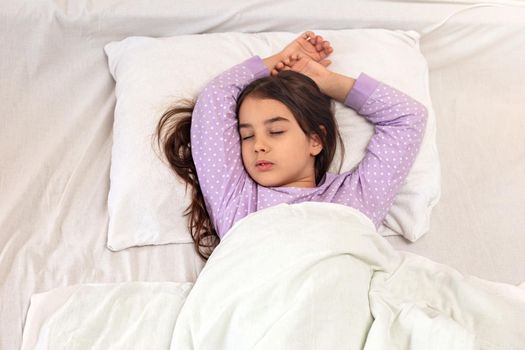 Little charming brunette girl with long hair in purple pajamas, sleeps lying on a white pillow, under a white blanket on the bed. View from above. Top view. Copy space