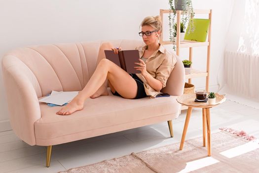 A modern woman of 30-40 years old, in casual clothes and glasses, sits on a comfortable beige sofa in a bright interior, with a diary and documents. copy space