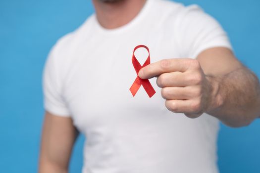Man with red ribbon bow AIDS awareness symbol in hand wearing a white t-shirt isolated on a blue background. Modern medicine and healthcare. AIDS awareness concept. No face visible.