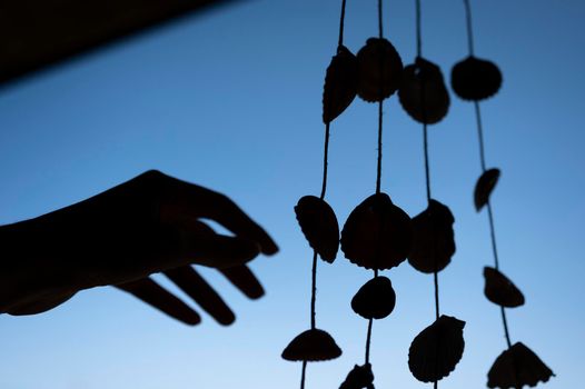 Silhouette of hand touching a dream catcher made with seashells against blue sky.