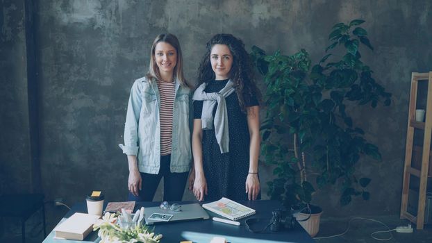 Two female business colleagues are standing together in loft style office, posing and looking at camera. Girls are happy, smiling and laughing