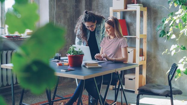 Young owners of small business are working with laptop in modern loft style office. Blonde is sitting and typing, brunette is standing near the desk and suggesting ideas looking at screen.