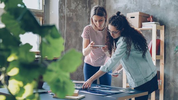 Pretty young women are standing near table, placing colorful pictures on it and shooting flat lay with smartphone. They are sharing ideas about design and smiling. Creative teamwork concept.