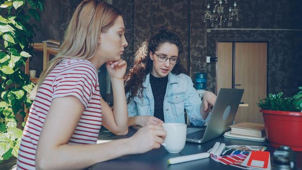 Attractive female coworkers are working at project together watching laptop screen. They are talking and gesturing, one of them is holding tea cup. Team brainstorming concept.