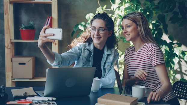 Young enterpreneurs are talking and making selfie with smart phone while sitting at desk in modern office. They are posing and laughing