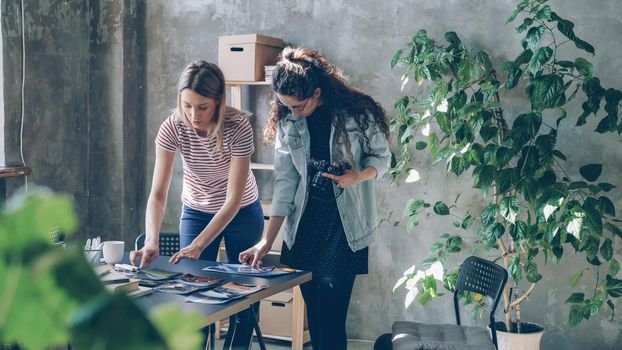 Young female enterpreneurs are placing photos on table to make flat lay and shooting with camera while standing in modern light office. Women are sharing ideas and discussing design.