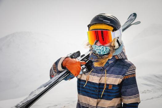 Portrait of a pretty and active woman skier, wearing a mask and holding skis in her hands, active winter holidays. extreme lifestyle concept