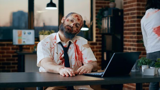 Bizarre looking zombie using modern laptop in office workspace. Evil apocalyptic brain-eating monster with deep and bloody wounds and scars trying to use portable computer at work.