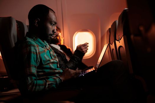 African american freelancer working on laptop during sunset flight, travelling to international destination to go on work trip. Young man flying in economy class with airplane transport.