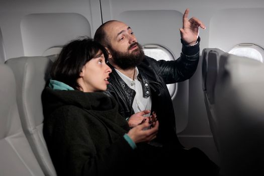 Couple of tourists waiting for flight to takeoff on airplane, being seated in economy class. Man asking flight attendant to help, flying with international airways plane to travel on holiday vacation.