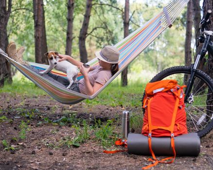 Caucasian woman lies in a hammock with Jack Russell Terrier dog in a pine forest.