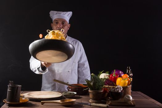 Male chef in white uniform holding a frying pan, sautéing spaghetti with fresh vegetables flying in the air before serving while working in a restaurant kitchen