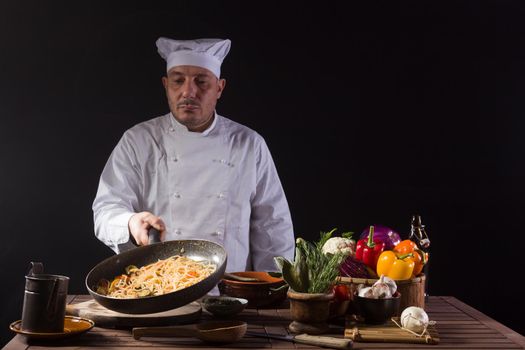 Male chef in white uniform holding a frying pan, sautéing spaghetti with fresh vegetables flying in the air before serving while working in a restaurant kitchen
